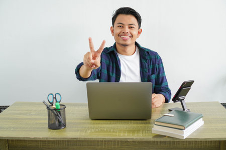 asian man sitting in workplace gesturing peace sign with fingers. Happy guy enjoying remote work at...