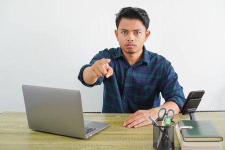 strict young asian man sit work at wooden desk with pc laptop. Achievement business career lifestyle concept. pointing index finger on cameraの写真素材