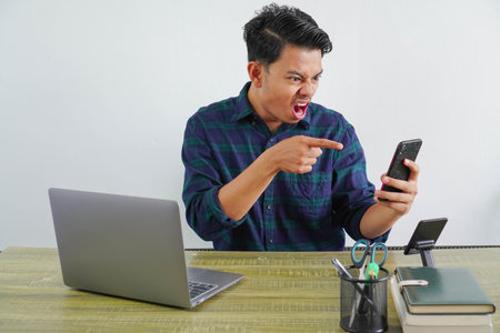 Angry young asian man sit work at wooden desk with pc laptop. Achievement business career lifestyle concept. talking and pointing on mobile phoneの写真素材