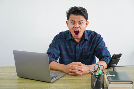 Angry young asian man sit work at wooden desk with pc laptop. Achievement business career lifestyle concept. screaming, swearingの写真素材