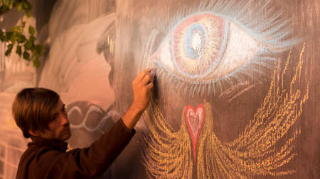 Side view of millenial man is drawing an eye and heart with wings on an outdoor chalkboard displayのeditorial素材