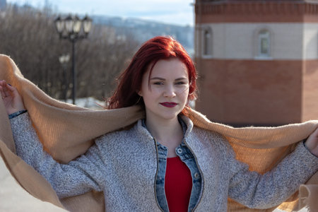 Pretty young women with red hair in beautiful beige scarf and grey coat posing in park.