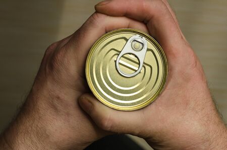 Two Hands of an Adult Man holds a can of canned food. Closed tin can with open key in hand. Canned foods. This really canned product. Selective focus. View from above. Tinted photoの写真素材