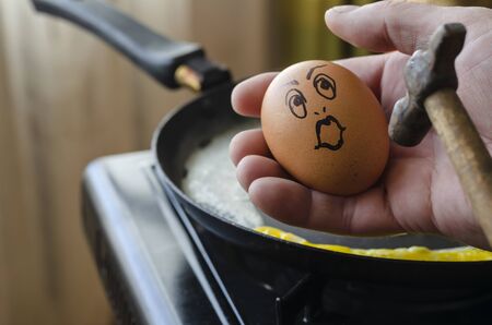 Surprised chicken egg in hand in front of a hammer. Raw chicken egg with a grimace of surprise over a frying pan with a scrambled egg. Never give up. Angled side view. Close-up. Selective focus.の写真素材