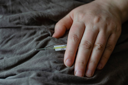Female hand lies on a blanket. In his hand is a mercury thermometer. The concept is bed rest,...