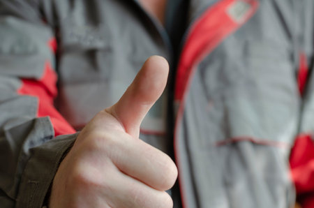 A man shows the camera a thumb up gesture. Mechanic in a red-gray working uniform with a raised...
