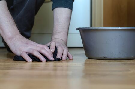 A man washes a floor with a rag. Adult man is manually washing the floors in the house. A gray plastic basin with water in the background. Homemade routine. Selective focus.の写真素材