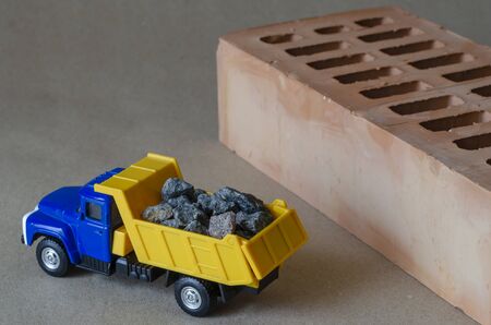A yellow dump truck carries rubble next to a brick. Toy truck with a full body of building materials. Creative industrial background. Construction and transportation of building materials. Selective focus.の写真素材