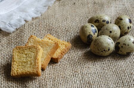 Square croutons and raw quail eggs on burlap. Three small croutons and a group of eggs. Still life in a rural style. Top view at an angle. Selective focusの写真素材