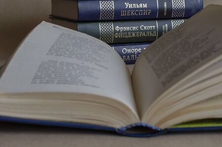 An open book on a table in a library. In the background is a stack of hardback books. Inscriptions on the roots of books: William Shakespeare, tragedy, F.S. Fitzgerald, the great Gatsby, Honore de Balzacの写真素材