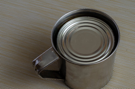 Steel mug and tin can on a textured background. A can of canned food in a stainless steel mug....