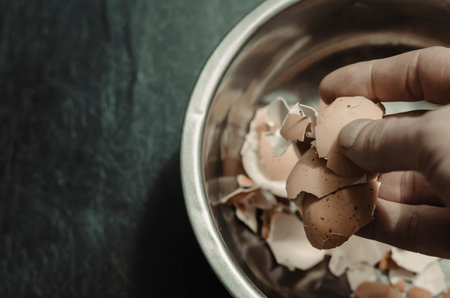 A male hand dips eggshells in a metal bowl. A metal bowl with eggshell on a black background. Adult...