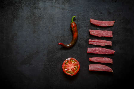 Flat lay Raw beef strips, half ripe tomato and hot pepper pod on black. Serving of raw beef fillet and raw vegetables.の写真素材