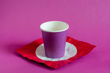 Creative background with lilac paper cup and colored foil. Paper cup for coffee on a stand made of red foil with a white circle. Side view at an angle. Selective focus.の写真素材