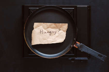 Flat lay scrap of brown paper with single word Hungry on a frying pan. Handwritten text on a torn piece of paper. The frying pan is on the gas stove. Recipe, menu, cooking concept.の写真素材