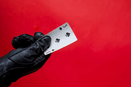 Male hand in leather glove holds playing card. The right hand in black glove with triplet of clubs on corral background. Fortune telling or magician concept. Side view. Selective focus.の写真素材