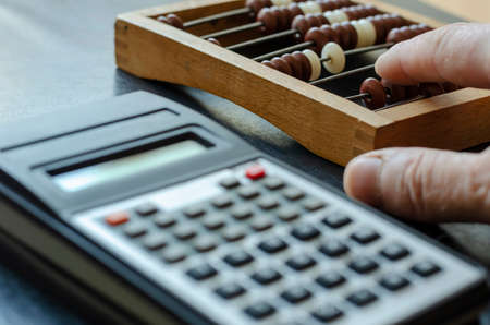 A middle-aged Caucasian man makes calculations on a wooden abacus. The calculator is nearby. Close-up of a male hand with an obsolete device for mathematical calculations.の写真素材