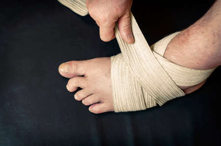 Middle-aged Caucasian man produces an independent bandage of his dislocated leg. He puts an elastic bandage over his swollen shin. First aid in sprains and dislocations. Bandage stabilizes ankle joint.の写真素材