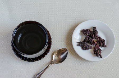 Black glass cup with boiling water, metal spoon and dry hibiscus petals. Dehydrated Sudanese rose...