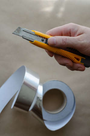 Middle-aged Caucasian man hand holds an orange knife. Roll of foil tape in the background. Heat and moisture insulation services. Modern insulating materials. Selective focus.の写真素材