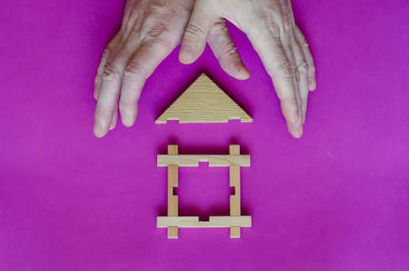 Hands are protecting a small toy wooden house. The hands of an adult man protect a wooden house on a purple background. Real estate insurance. Private property protection.の写真素材