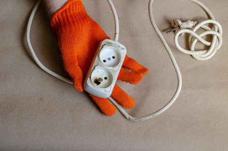 Hand wearing a protective orange glove holds an electrical outlet after fire. White power strip after short circuit. Electrical safety.の写真素材