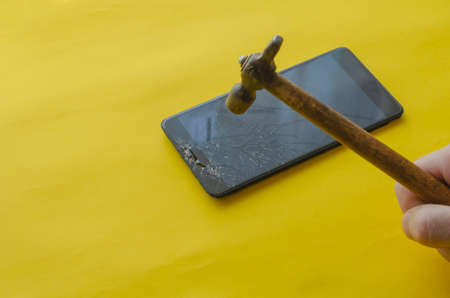 Broken black smartphone and small hammer on a yellow background. The hand breaks the display of the smartphone. Treatment of nomophobia or phone addiction. Selective focus.の写真素材