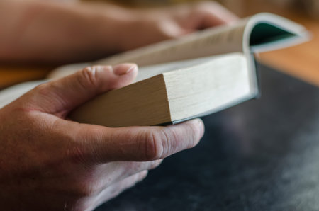 Male hands are holding an open book. A mature man is reading a thick hardback book on a black table....