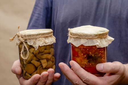 Male hands are holding glass jars of canned food. Transparent jar with lecho and mushrooms. Canned...