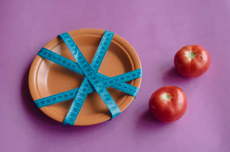 Fresh tomatoes on purple background and plate tied with measuring tape. Two ripe tomatoes and clay plate tied crosswise with measuring tape. Overeating control, diet, nutritionist.の写真素材