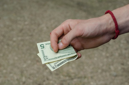 A man's hand holds a 5 American dollar bill. Right hand with a red braided bracelet. Charity and...