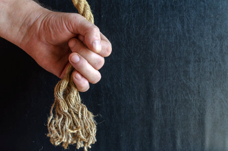 A man's hand holds a rope with a knotted knot on a dark background. A mature man with a thick rope...