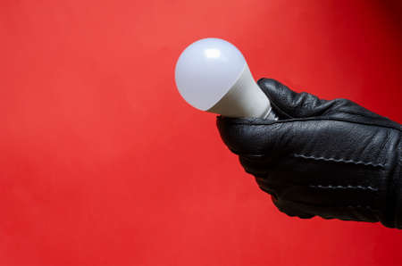 Male hand in leather glove holds light bulb. Right hand holding single white light bulb against coral background. Side view. Selective focus.の写真素材