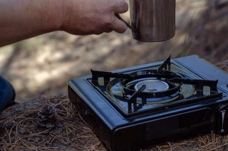 Herbal tea in a metal mug in hand over a portable gas stove. A man's hand holds herbal tea made from rose hips and various herbs in a stainless steel cup. Hiking tourism concept. Selective focus.の写真素材