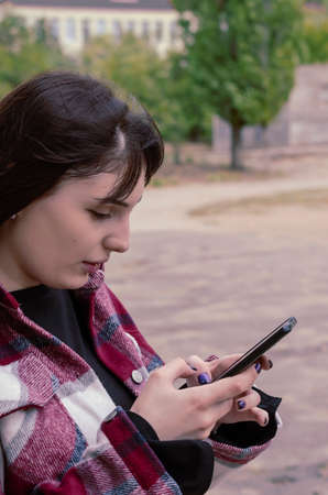 Portrait of a young woman writing a message on the phone. Woman with long flowing hair in a plaid shirt outdoors. Lifestyleの写真素材