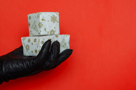 A hand in a black leather glove holds a wrapped Christmas present on a red background. Small...