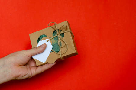 Cardboard box with gift and hand on red background. Man's hand holds wrapped gift box. Brown cardboard box tied with twine.の写真素材