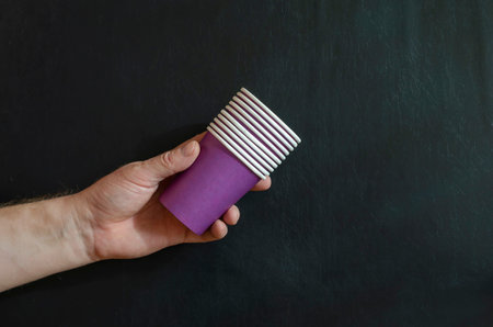 A man's hand holds a stack of lilac coffee glasses on a black background. Paper cups for hot drinks....