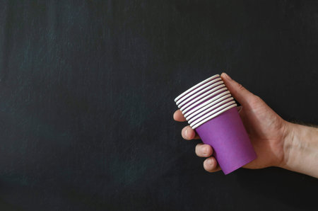 A man's hand holds a stack of lilac coffee glasses on a black background. Paper cups for hot drinks....