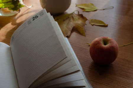 An open diary, ripe apple and fallen leaves on the desk. Autumn creative background without anyone. Selective focus.の写真素材