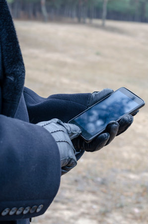 Businessman in black coat and black leather gloves holds the phone in his hand as he walks through the park. Close-up, selective focus.の写真素材