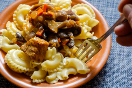 Defocused, soft focus, selective focus, blur. A man's hand pricks pasta in a clay plate with a fork. Ready-to-eat pasta with stewed vegetables, meat and mushrooms.の写真素材