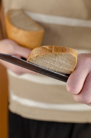 A man's hand with a knife in it holds out a freshly cut loaf of bread to the camera. Sharing food,...