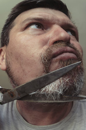 An adult man cuts his own beard with old huge scissors. Unusual portrait of a bearded man with gray...
