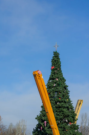 Dismantling the city's main Christmas tree. Yellow booms Two mobile cranes and a tall Christmas tree with ornaments on the background of the blue sky. No people.の写真素材