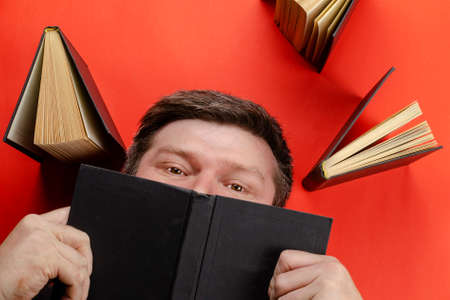 The benefits of reading books concept. A middle-aged man with gray hair covered his face with a book. Red background. Flat layout.の写真素材