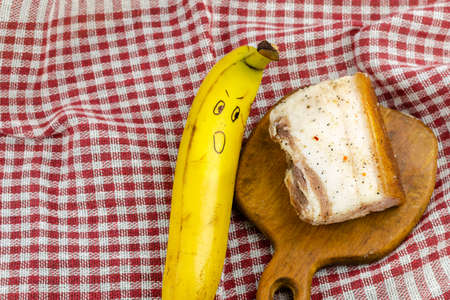 Banana with a smiley face and a piece of salty fat on a white and red checkered cloth. Ripe banana with a grimace of surprise. Salty fat on a wooden cutting board. An angled view from above. Selective focus.の写真素材