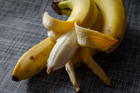 Three unpeeled bananas, on top of which lies the fourth, with a partially peeled peel. Ripe bananas on a gray surface. Focus on the foreground.の写真素材