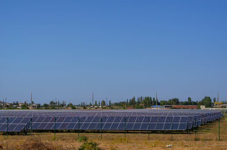 Rows of solar panels behind a fence in a field. A solar power plant against the backdrop of a small...