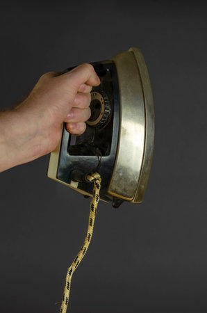 Man's Hand holds old rusty iron on a gray background. Old metal iron for ironing with an electric wire. Selective focus.の写真素材